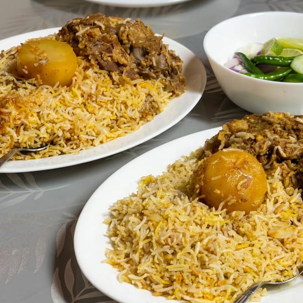 Two plates of spiced rice with potatoes and braised meat, served alongside a bowl of sliced cucumbers, chilies, onions, and lime.