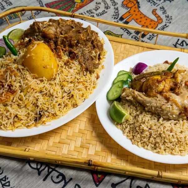 Two plates of spiced rice with chicken, potatoes, cucumber, lime, and fried onions on a bamboo tray.