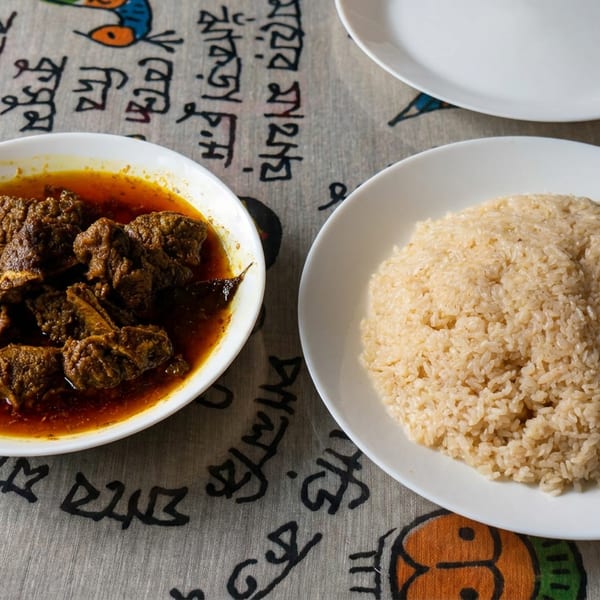 Bowl of richly spiced meat curry beside a mound of rice on white plates over a colorful patterned tablecloth.